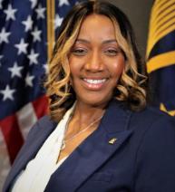 African American woman in a white top and blue blazer poses in front of the U.S. and Senior Executive flags