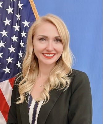 amanda french with blonde hair black suit jacket striped blouse posing in front of american flag 