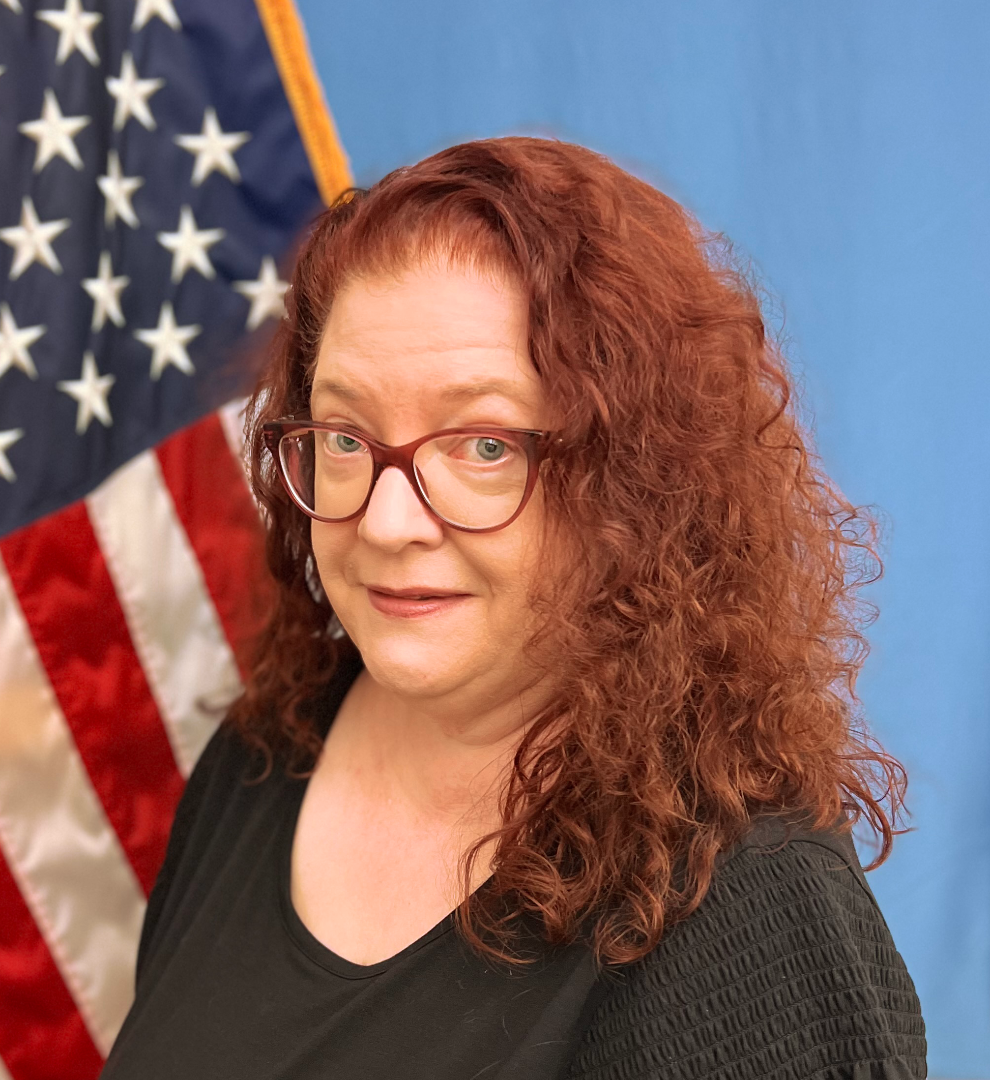 jill rufener with curly red hair with glasses and black blouse posing in front of american flag with blue background 