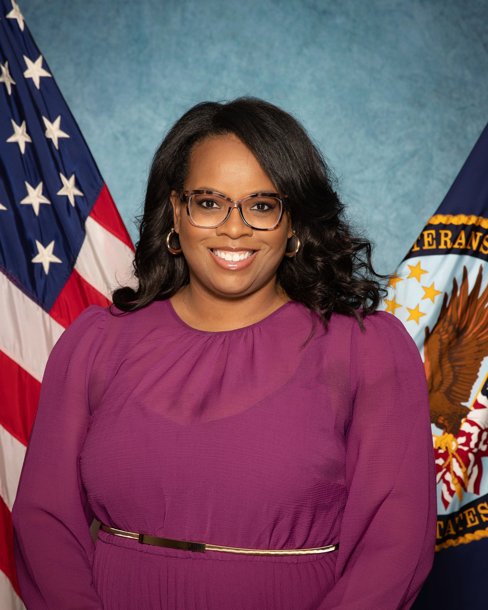 nikki wilder with long black hair and glasses and purple blouse posing in front of american flag and veterans affairs flag with blue background 