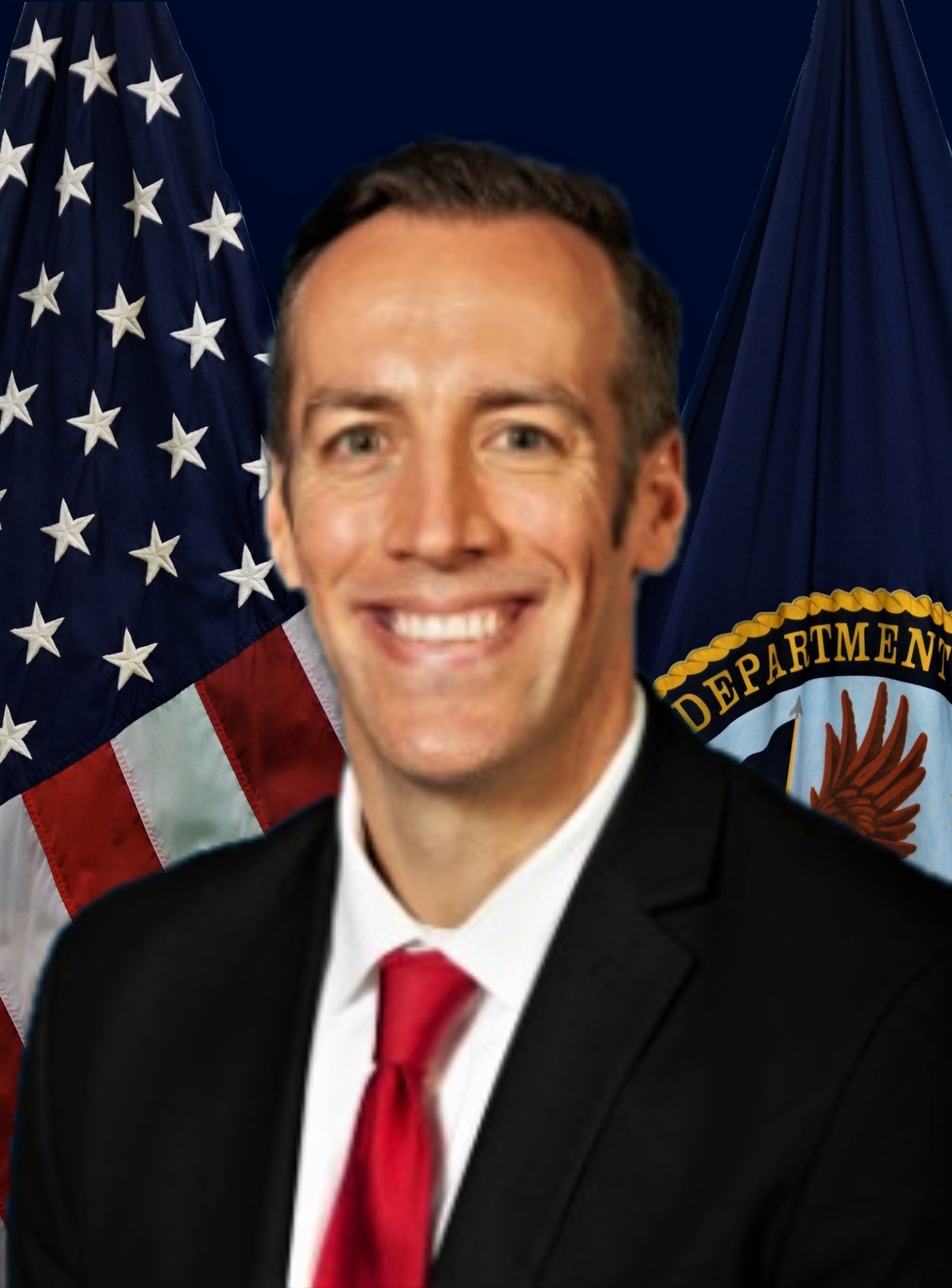 A man in a black jacket, white shirt, and red necktie poses in front of the United States and Department of Veterans Affairs flags.