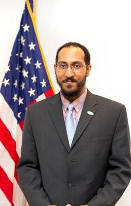 Male poses in grey blazer and light colored shirt in front of U.S. flag