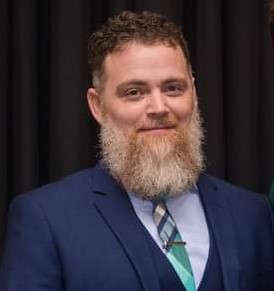 Male in a light brown blazer and blue shirt poses in front of blue background