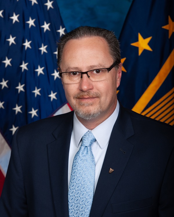 Mark poses in front of United States flag and Selective Executive Service flags wearing a dark-colored suite and glasses