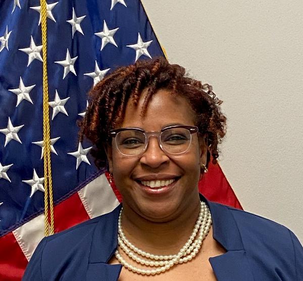 Craleta poses in front of United States flag wearing a dark-colored jacket, pearls and glasses
